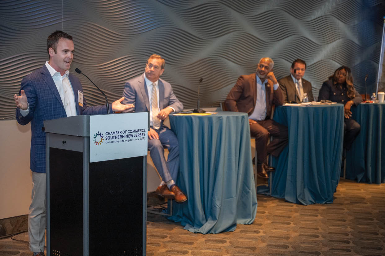 A person in a blazer is speaking at a podium labeled Chamber of Commerce Southern New Jersey while four people in business attire sit at tables covered with blue cloths in the background. Description generated by AI