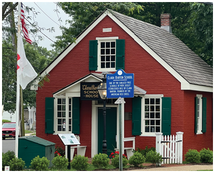 A small red brick building with green shutters and a sign identifying it as Clara Barton School, located next to a white picket fence and flagpoles. Description generated by AI