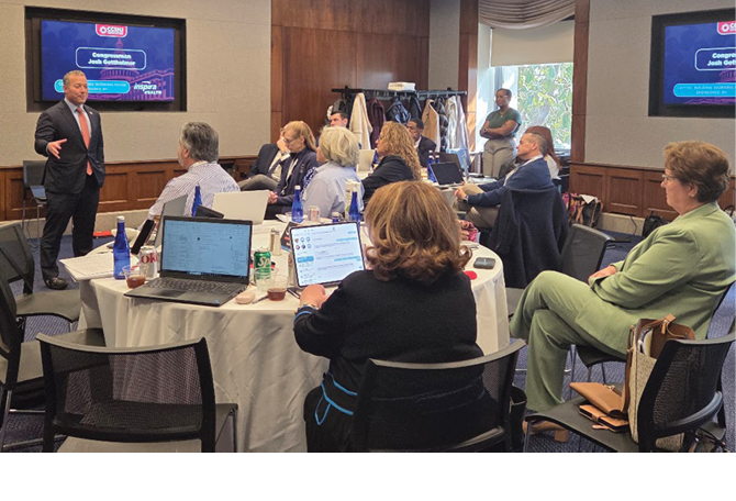 A group of adults seated around a table attentively listening to a person in a suit giving a presentation in a conference room with two screens displaying the same slide. Description generated by AI