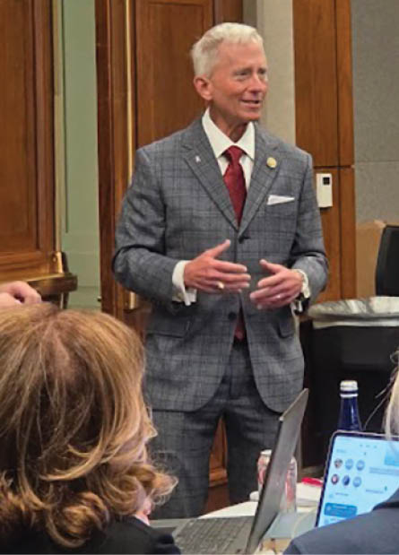 A person in a gray plaid suit and red tie is standing and speaking in a wood-paneled room while two people with laptops listen. Description generated by AI