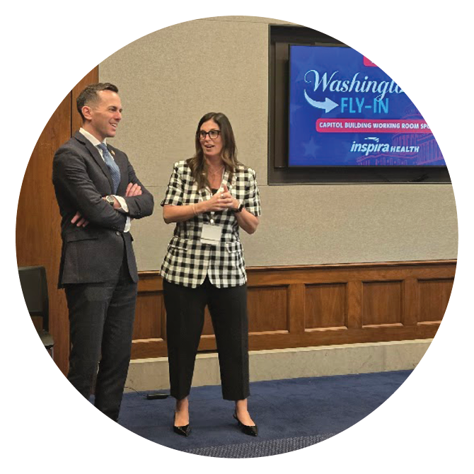 Two people stand in a room with wood paneling and a blue carpet, one wearing a checked blazer and the other in a dark suit, near a screen displaying information about a Washington Fly-In event. Description generated by AI