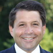 A person with neatly combed dark hair is wearing a dark suit jacket and white shirt, standing outdoors with greenery in the background. Description generated by AI