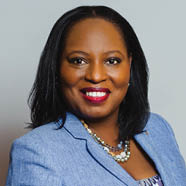 Person with shoulder-length straight hair wearing a light blue blazer, a patterned top, and a multi-strand beaded necklace against a plain background. Description generated by AI