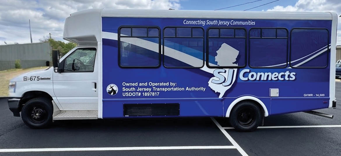 A blue and white shuttle bus with the logo “SJ Connects" and text indicating it is operated by the South Jersey Transportation Authority is parked in a lot under a partly cloudy sky. Description generated by AI