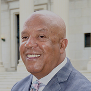 A close-up of a senior person with a bald head and a calm expression standing outdoors near a building. Description generated by AI