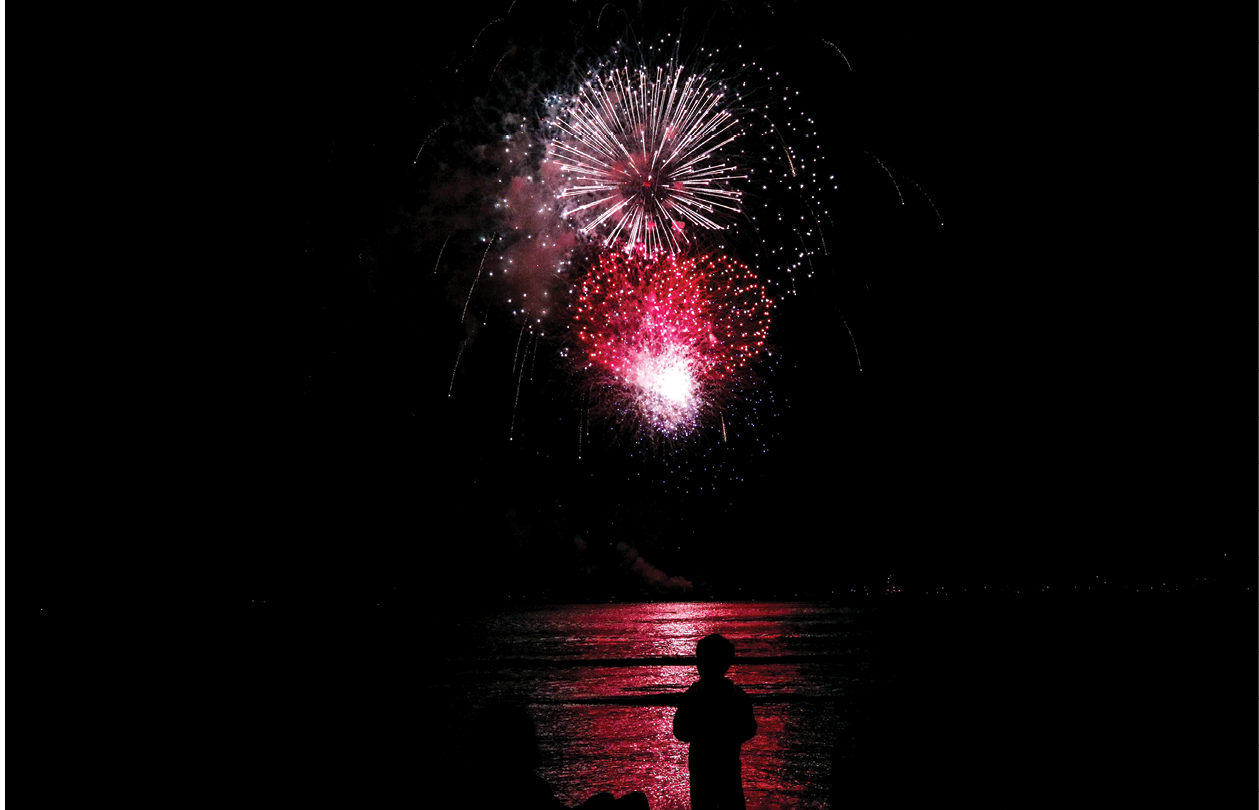 Brilliant fireworks explode in the sky, illuminating the water and creating a dazzling display of light and color. A silhouette of a person stands by the shore, capturing the awe of the spectacle.
