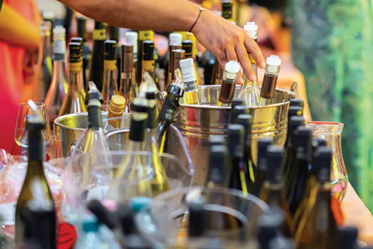 Hand reaching for wine bottle from metal ice bucket at festive wine tasting with many labels visible. Concept of wine selection, testing, celebration and premium alcohol