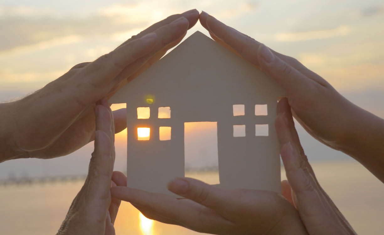 Group of human palms on all sides of a cut out house diagram with setting sun coming through window over water