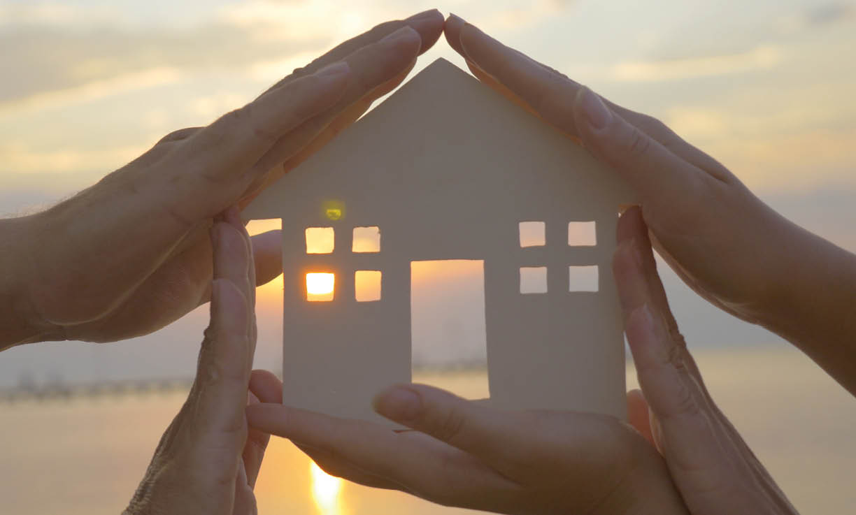 Group of human palms on all sides of a cut out house diagram with setting sun coming through window over water