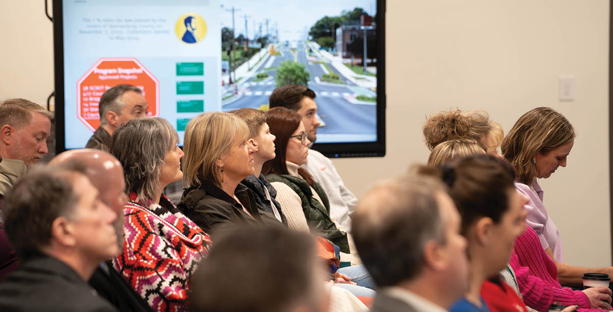 A group of people are sitting in a room, watching a presentation on a large screen. The presentation is about a road project. AI generated content