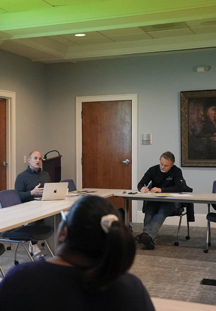 Three people are sitting around a table in a classroom setting. One man is using a laptop, while the other two are engaged in conversation. The room has a projector screen and a painting on the wall. AI generated content