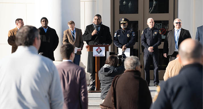 A group of people are gathered outside a building, with a man speaking into a microphone. There are several police officers standing nearby, some of whom are wearing ties. The crowd appears to be listening attentively to the speaker. AI generated content