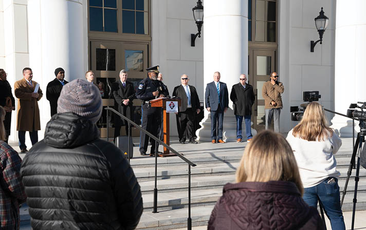 A group of people are gathered outside a building, with some of them wearing ties. A man in a suit is speaking into a microphone, while others stand around him. A woman is filming the event with a camera. AI generated content