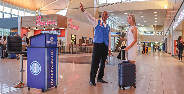 A man and woman are standing in an airport, the man is pointing upwards. The woman is holding a suitcase. AI generated content
