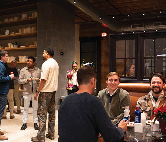 A group of people are gathered in a room, some sitting at a table and others standing. They are holding drinks and engaging in conversation. A man is sitting at the table holding a cell phone. AI generated content