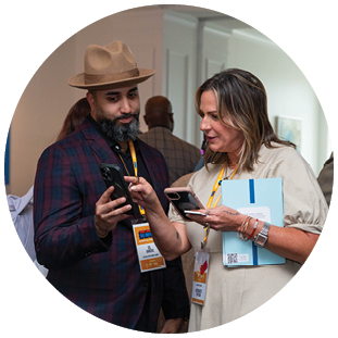 A group of people are gathered in a room, with some of them looking at their cell phones. A man in a plaid shirt is checking his phone, while a woman in a white dress stands nearby. There are also other people in the room, some of whom are wearing name tags. The scene appears to be a social gathering or event. AI generated content