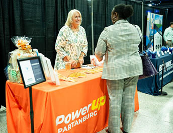 Two women are standing at a table with a sign that says “Power Up Partnership." One woman is wearing a suit and the other is wearing a dress. They are talking to each other. AI generated content