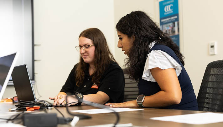 A group of people are sitting in a room, some of them wearing name tags. They are looking at their cell phones, possibly checking messages or browsing the internet. There are several cups and bottles on the tables in front of them, suggesting that they might be attending a meeting or event. AI generated content