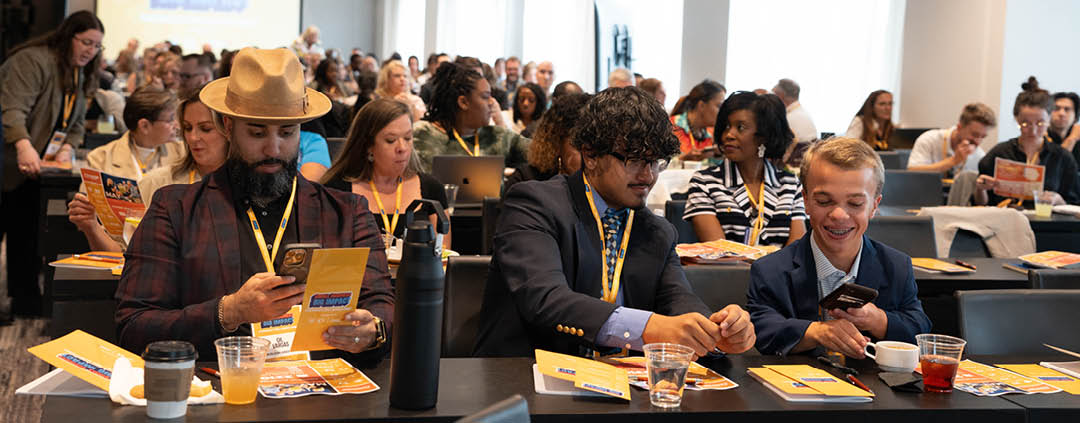 A group of people are sitting in a conference room, some of them wearing name tags. They are engaged in a presentation, with a TV screen visible in the background. The attendees are seated at tables, with cups and bottles placed on them. There are also laptops and cell phones in the room, indicating that the attendees are using them for note-taking or communication. AI generated content