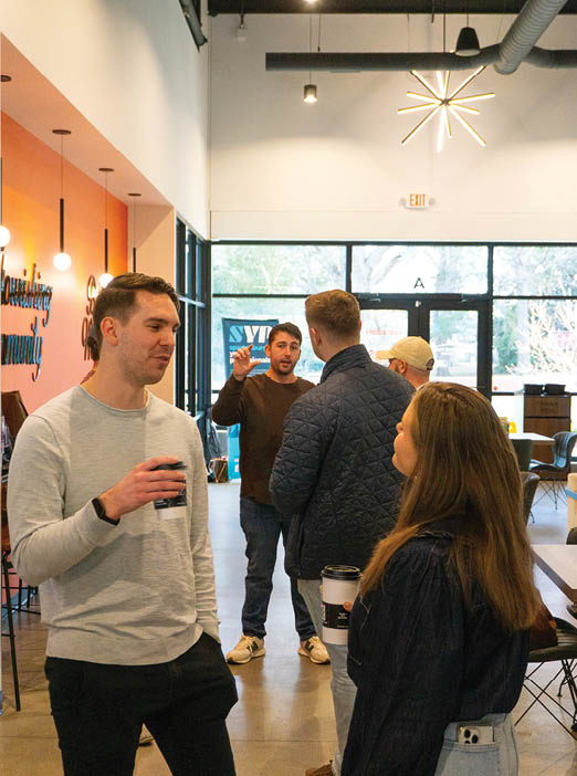 A group of people are standing in a room, engaged in conversation. One man is holding a coffee cup. AI generated content