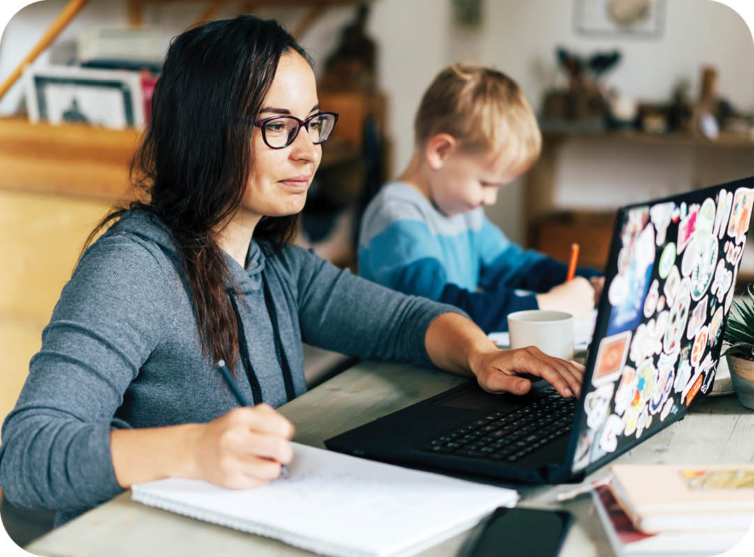 Concept of work from home and home family education. Mom and son are sitting at the desk. Business woman works on the Internet in a laptop, a child writes in a notebook.