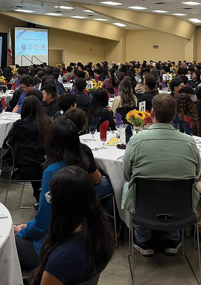 A large group of people are sitting in a room, attending a conference. They are seated at tables with plates, forks, knives, and spoons. There are also chairs and dining tables in the room. The attendees are engaged in the event, and there is a TV screen visible in the background. AI generated content