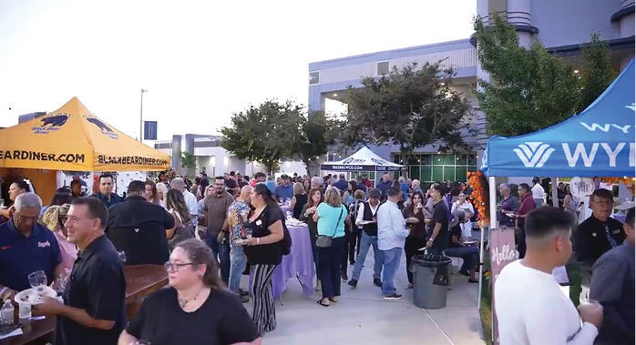 A large crowd of people are gathered in a parking lot, with many of them holding wine glasses. There are several tents set up, and a building can be seen in the background. AI generated content