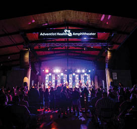 A large group of people are gathered in a stadium, watching a concert. The performers are on stage, and the audience is seated in chairs surrounding the stage. The event is sponsored by Adventist Health Amphitheater. AI generated content