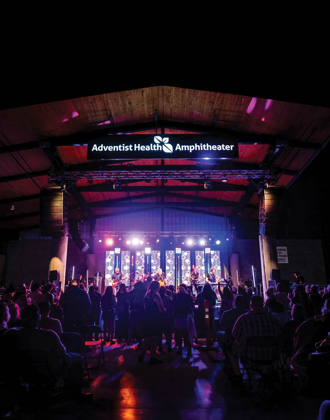 A large group of people are gathered in a stadium, watching a concert. The performers are on stage, and the audience is seated in chairs surrounding the stage. The event is sponsored by Adventist Health Amphitheater. AI generated content