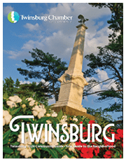 A tall stone monument with an eagle on top stands among trees and flowers under a partly cloudy sky with the text Twinsburg Chamber of Commerce and Twinsburg 2026 Community Guide Your Guide to the Neighborhood. Description generated by AI