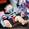 Baby sleeping in stroller, which is festively decorated with red, white and blue for the Fourth of July holiday. The baby is barefoot, has a blue pacifier and is dressed in red, white and blue.