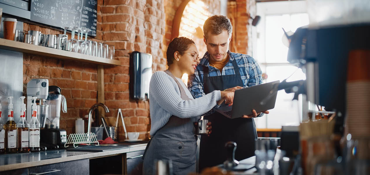 Two Diverse Entrepreneurs Have a Team Meeting in Their Stylish Coffee Shop. Barista and Cafe Owner Discuss Work Schedule and Menu on Laptop Computer. Multiethnic Female and Male Restaurant Employees.
