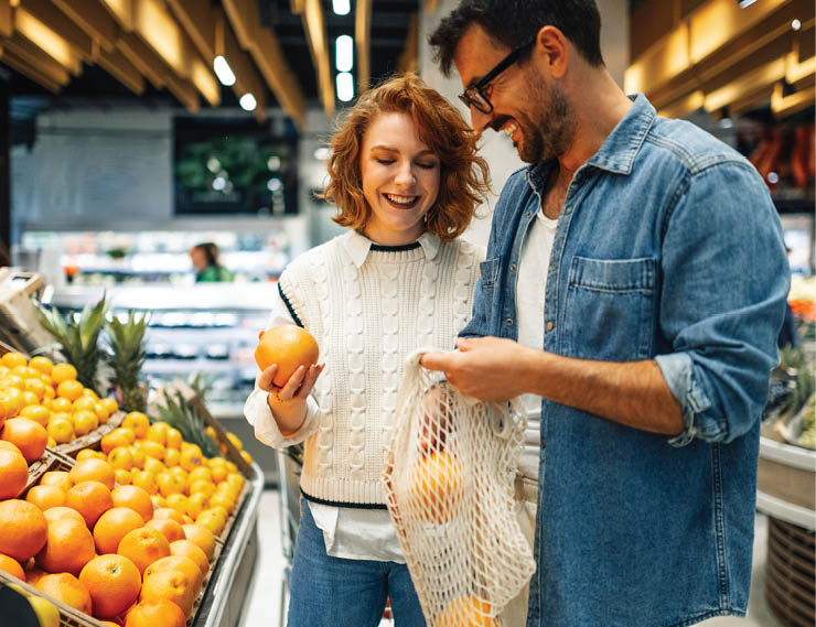 Young smiling couple buying fresh oranges at supermarket, holding reusable mesh bag, promoting sustainable and eco friendly lifestyle, choosing healthy food