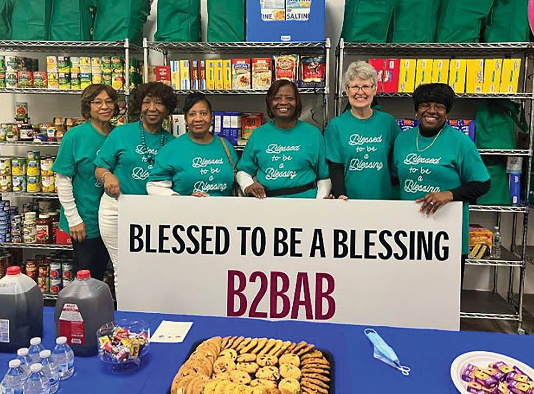 A group of women wearing green shirts are standing in front of a table with a sign that says “Blessed to be a blessing." Description generated by AI