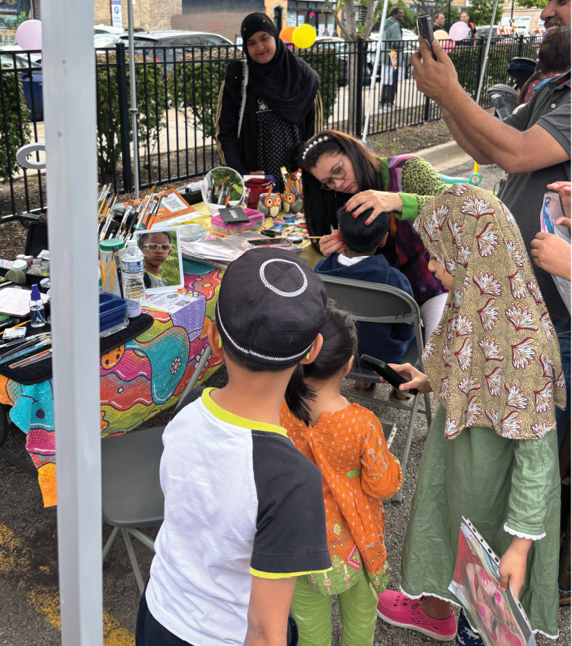 A group of people, including children, are gathered around a table with a woman painting. They are standing under a tent, and there are cars parked nearby. AI generated content