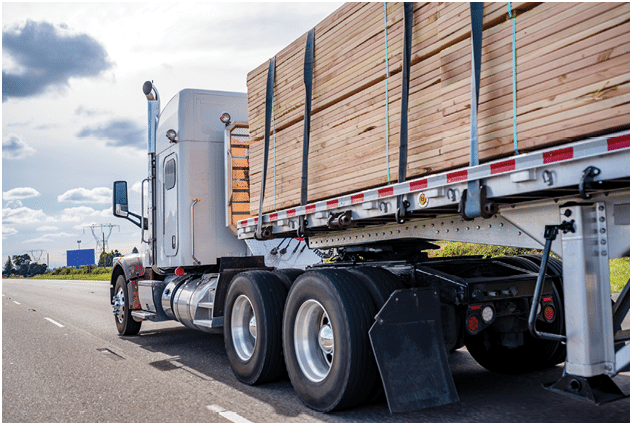 White classic powerful bonnet American big rig professional semi truck tractor transporting tightened wood lumber cargo on flat bed semi trailer running on wide multiline highway road