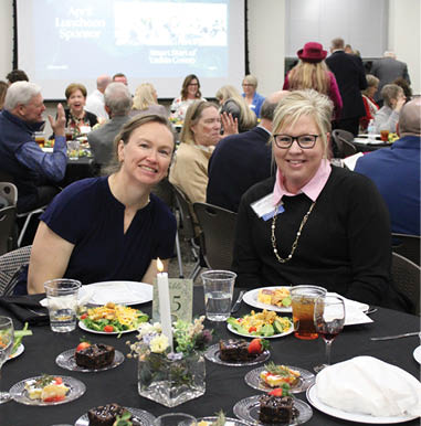 Two women are sitting at a table with plates of food in front of them. They are smiling and posing for a picture. The table is set with various items such as cups, forks, knives, and spoons. There are also other people sitting at tables in the background. AI generated content