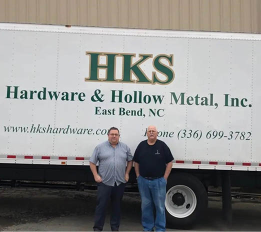 Two men are standing in front of a white truck that says “HK's Hardware & Hollow Metal Inc.". AI generated content