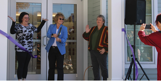 Three women are standing in front of a house, holding ribbons. One woman is wearing a blue jacket. AI generated content