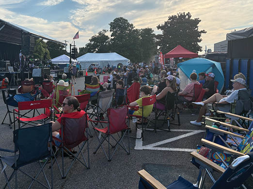 A large crowd of people are sitting in chairs on a street, watching a performance. There are several umbrellas providing shade for the audience. AI generated content