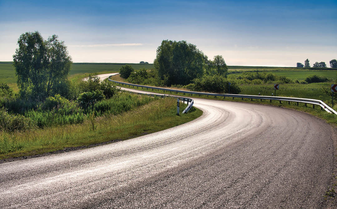 Empty, curved road is flanked by grassy fields with some small bushes, trees, and shrubs scattered around and guardrail extends along the road