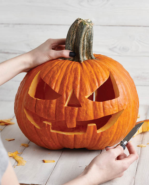 Woman carving big orange pumpkin into jack-o-lantern for Halloween holiday decoration on white wooden planks, close up view