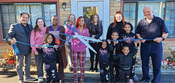 A group of people, including children, are standing in front of a house. They are holding scissors and a ribbon, preparing to cut it. AI generated content