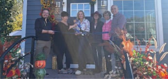 A group of people are standing in front of a house, posing for a picture. They are holding a ribbon and a pair of scissors, indicating that they are about to cut the ribbon. The people are of different ages and genders, and they are all smiling for the camera. AI generated content
