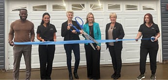 Five people are standing in front of a garage, holding a blue ribbon. They are posing for a picture. AI generated content