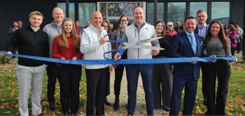 A group of people are standing in front of a building, posing for a picture. They are holding a blue ribbon, which is being cut by a man in a suit. The people are wearing ties, and some are holding cell phones. AI generated content