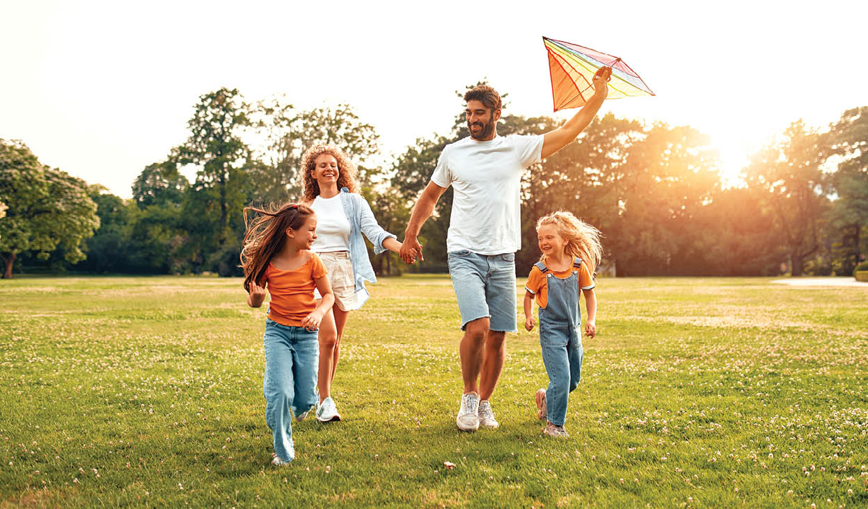 Happy young family dad, mom and two daughters running and playing with a colorful kite in a meadow in the park, having fun on a warm sunny weekend.