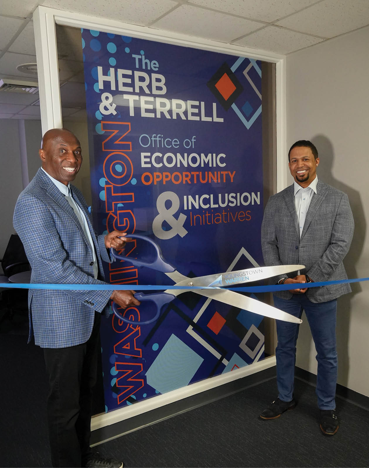 Two men cutting a blue ribbon in front of a blue and orange sign. Description generated by AI