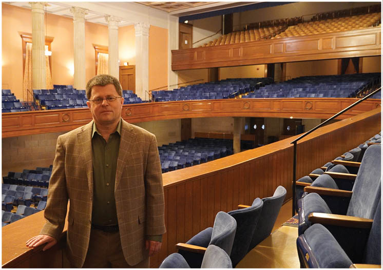 A man wearing a suit and tie standing in front of a large auditorium. Description generated by AI